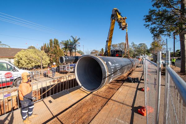 A large, black PVC water pipe is suspended from a crane and being pulled into position by two men, both dressed in high visibility protective clothing. The photo is taken on a residential street, and the work site is fenced off on either side.